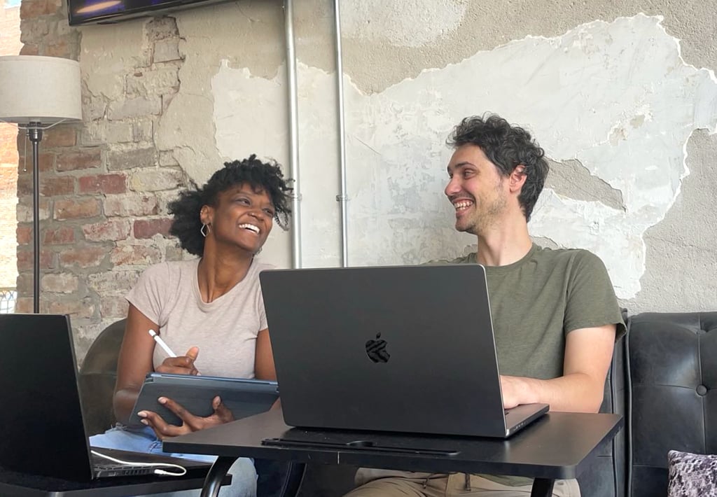 Black woman smiling to her male white friend, apple computer, notepad, brick wall