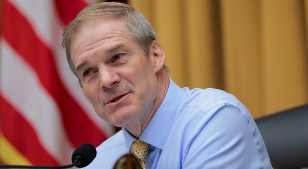 Jim Jordan, grey hair, blue shirt, mustard-color curtais, US flags' white and red stripes