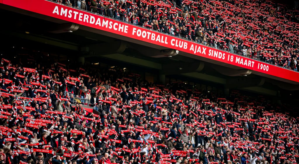 Supporters of AFC Ajax Amsterdam stand in stadium with club merch scarfs up.