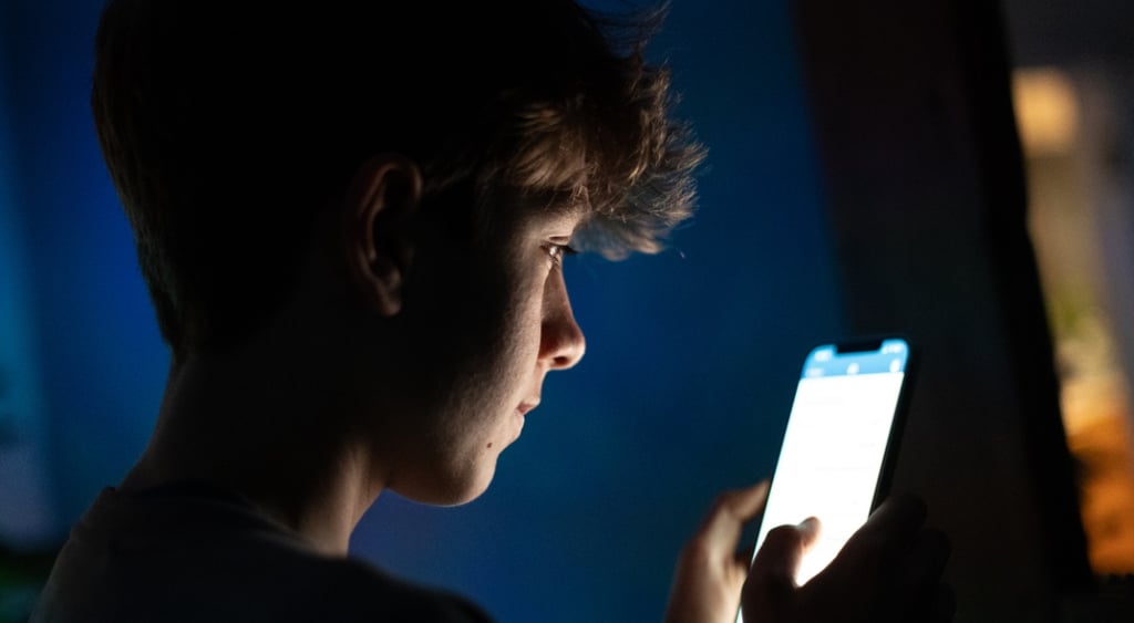 young teen boy scrolls phone in the dark, white flashy screen,