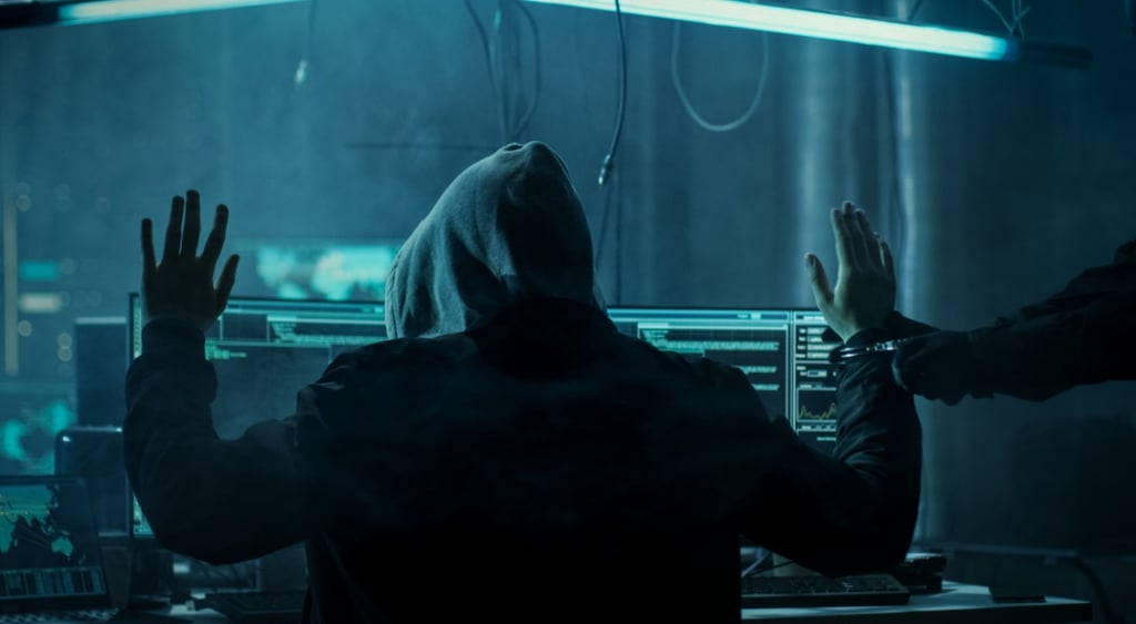 dark web hacker in hoodie, arms up, left arm handcuffed, arrest, dark blue light room