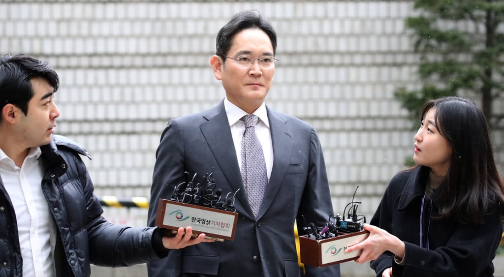 Lee Jae-yong in grey suit, white shirt, violet tie, woman in black suit, microphones, glasses