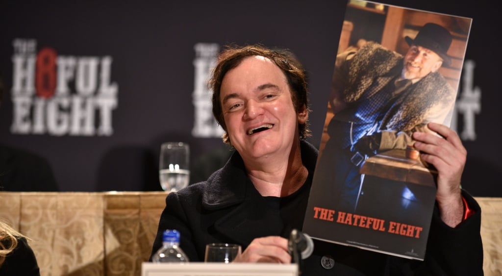 Quentin tarantino in blavc suit smiling, holding poster for the hateful eight