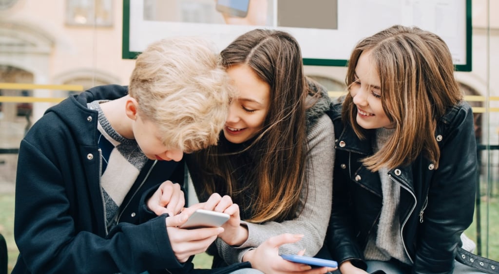 two teen girls, brunettes hold smartphones, one blonde teen boy, wait in bus stop, laugh