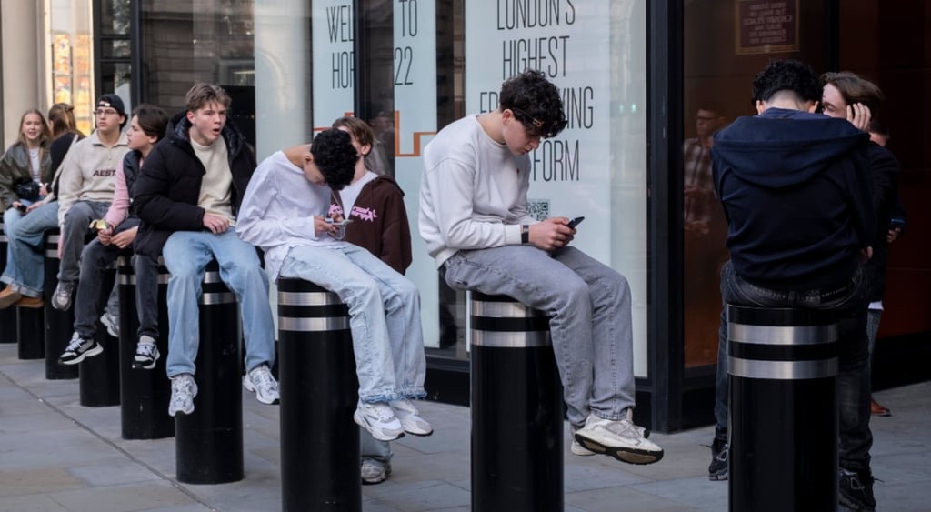 ten young people sit on bollards, scroll their phones, blue jeans, grey jumpers,