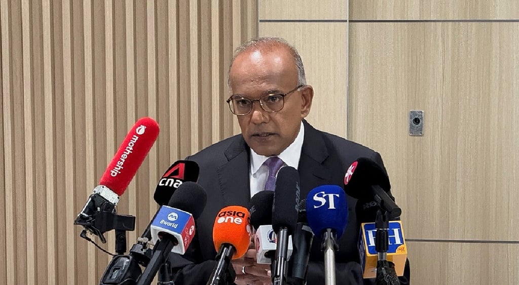K Shanmugam, brown skin, bald man, some grey hair, about ten microphones, wooden wall