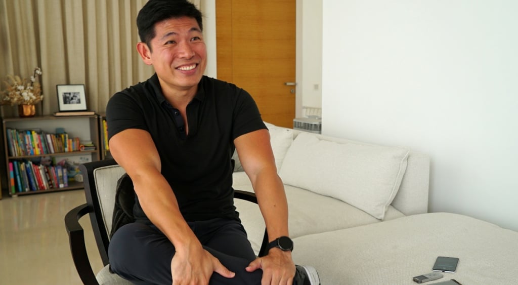 asian man from alaysia, smiling, black hair, black t-shirt, white sofa, white wall, hands on knees,