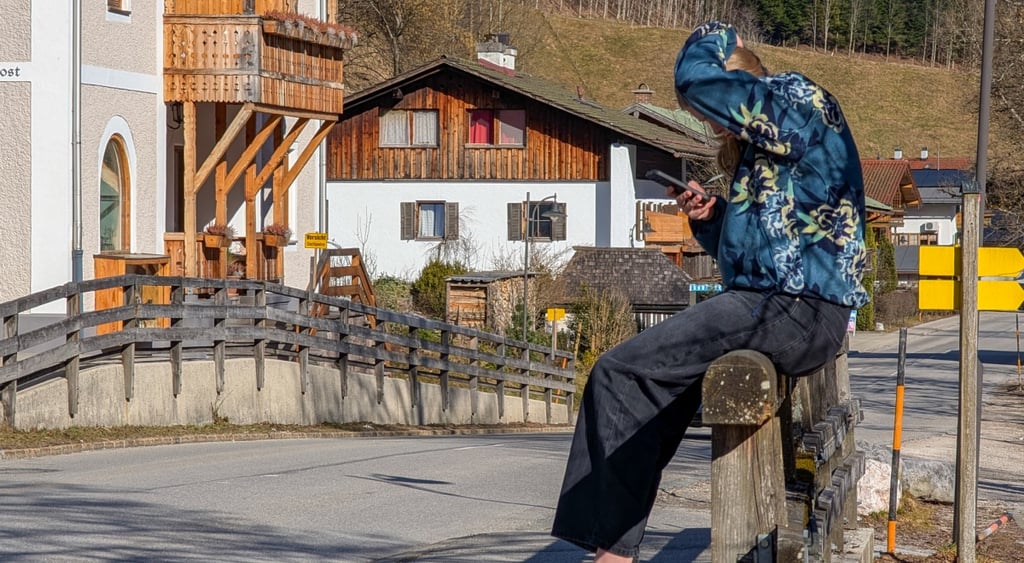 woman sits on wooden railing, looks at smartphone, wooden house, black jeans, blue hoody