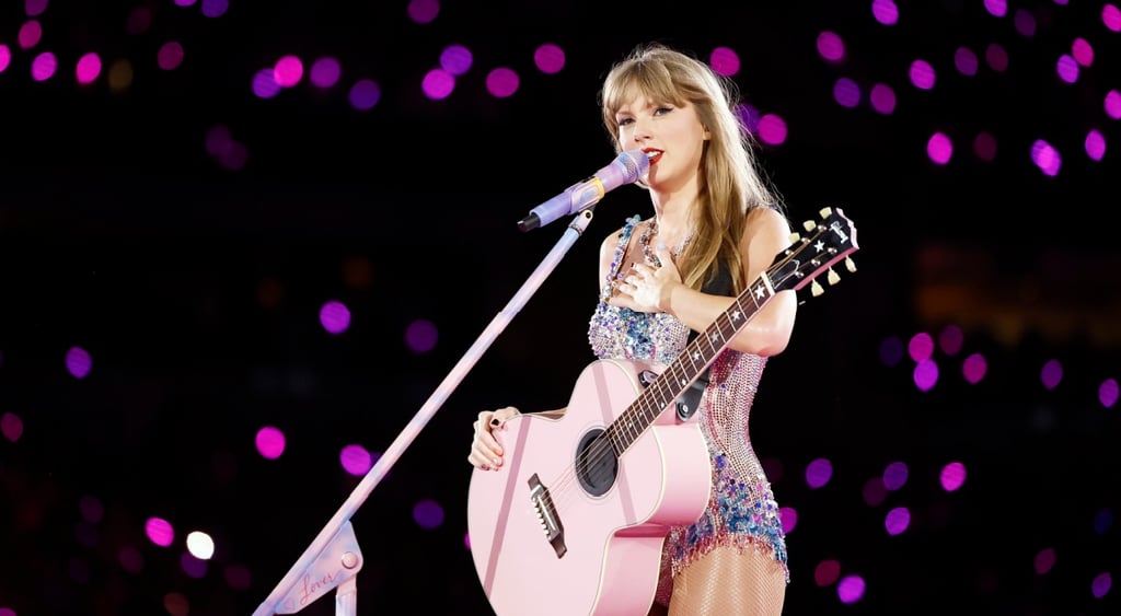 Taylor Swift, pink-violet sparkly bodysuit, pink guitar, white microphone, dark stadium crowd