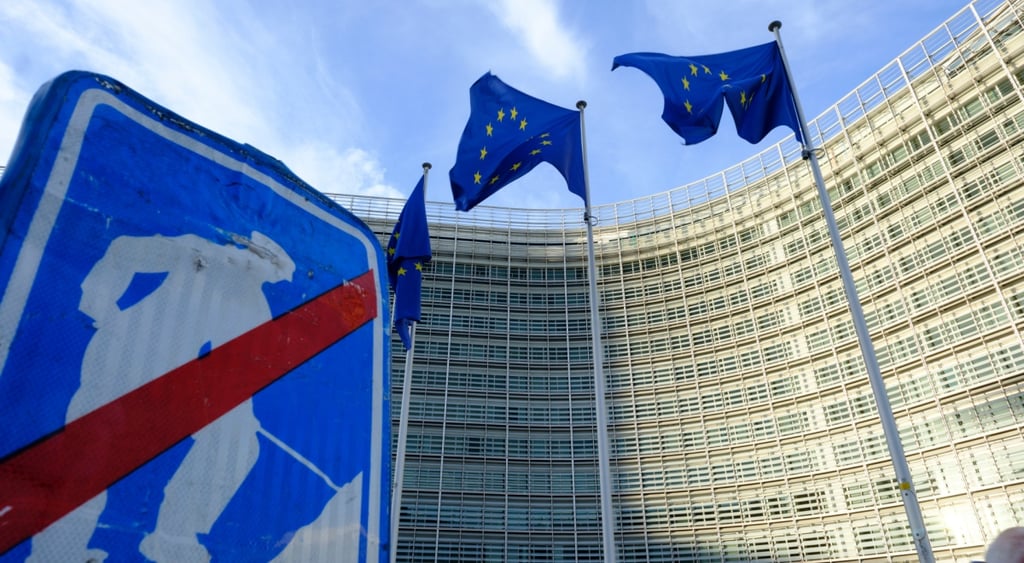 blue indication sign, white man figure digging, berlaymont building, sunny day, three eu flags