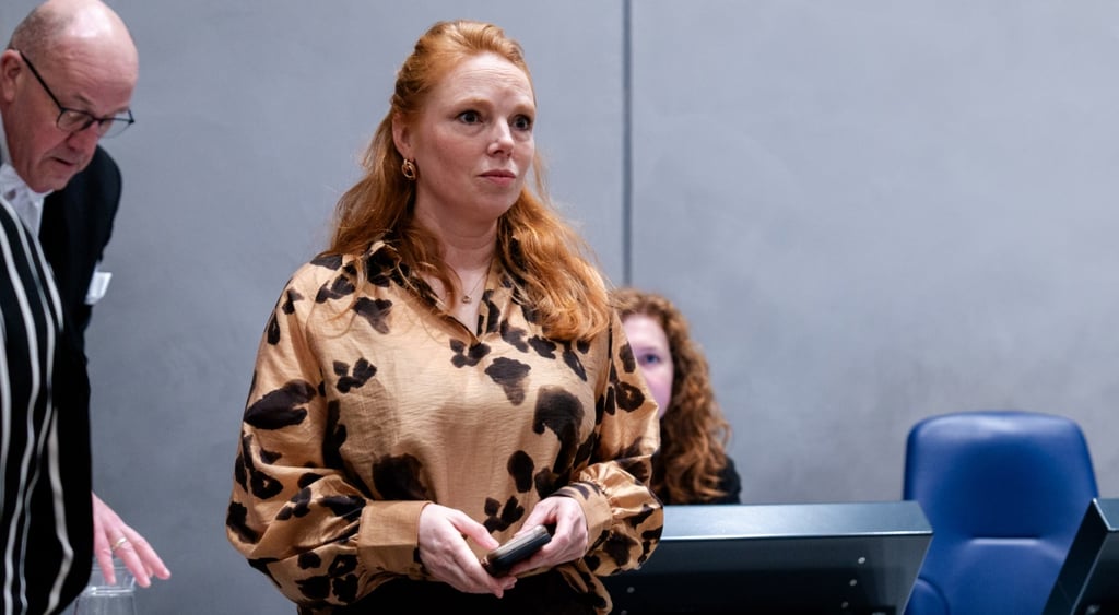 white woman in her 40s, long bright ginger hair, orange shirt with black dots, grey wall, blue chair
