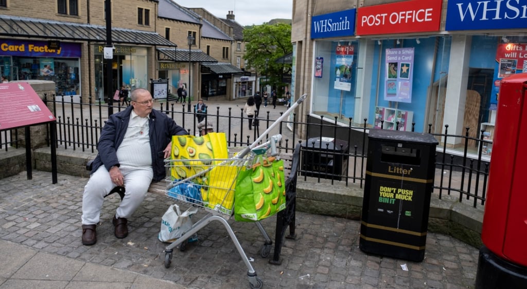 yellow asda shopping bad, man on bench, hallifax, grey pavement, grey sweatpants