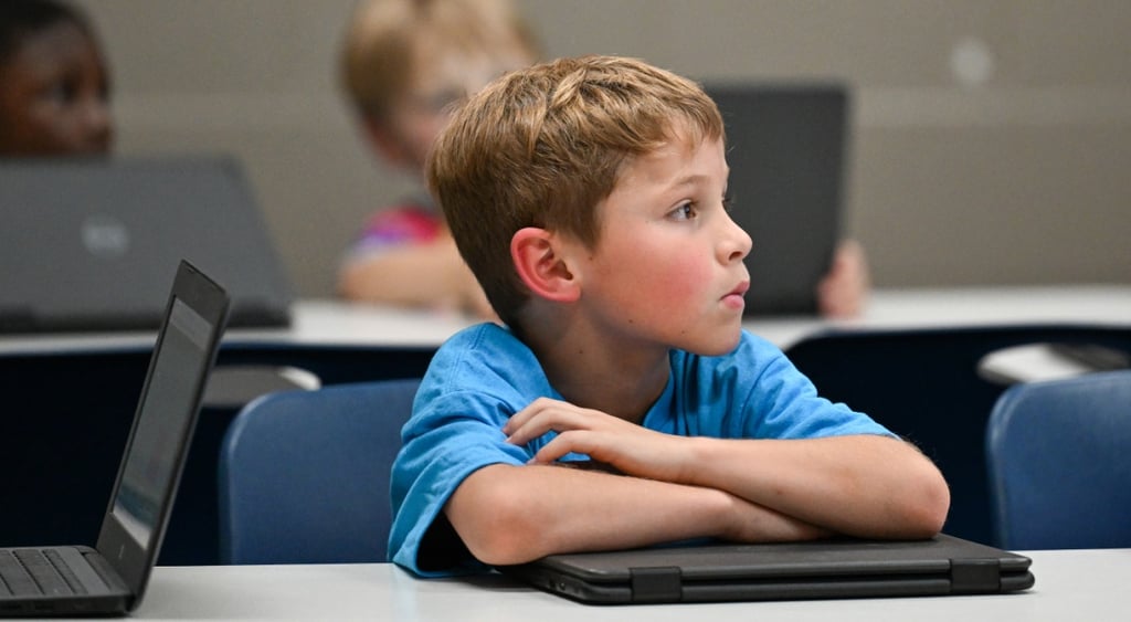 white blonde boy, white desk, hands on black laptop, open black laptop, blue t-shirt,