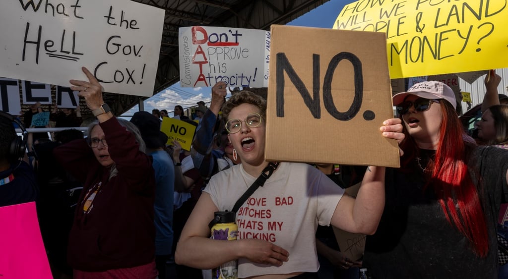 white woman, short curly blonde hair, protest signs, many people, yellow framed glasses