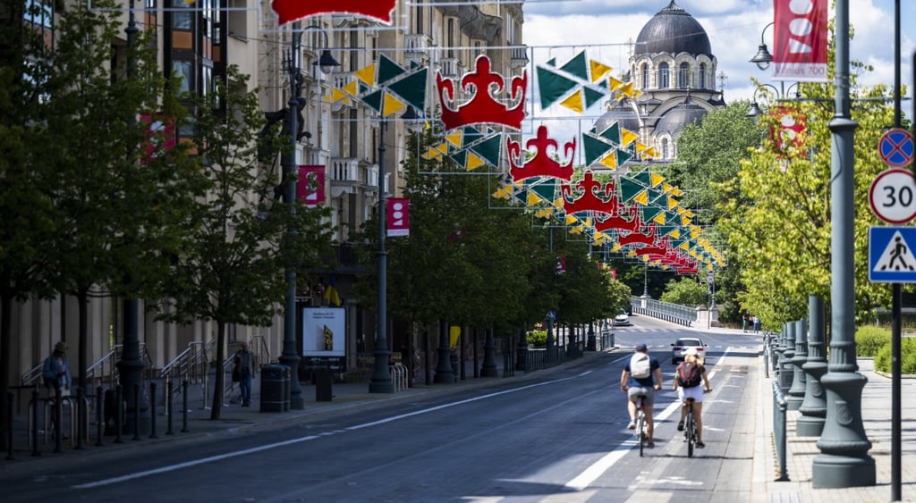 green trees, two cyclists, street, dark grey church tower, red flags, blue sky, white clouds