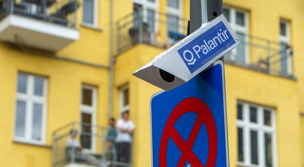 white surveillance camera on grey poll, blue sticker with palantir written on it, yellow building