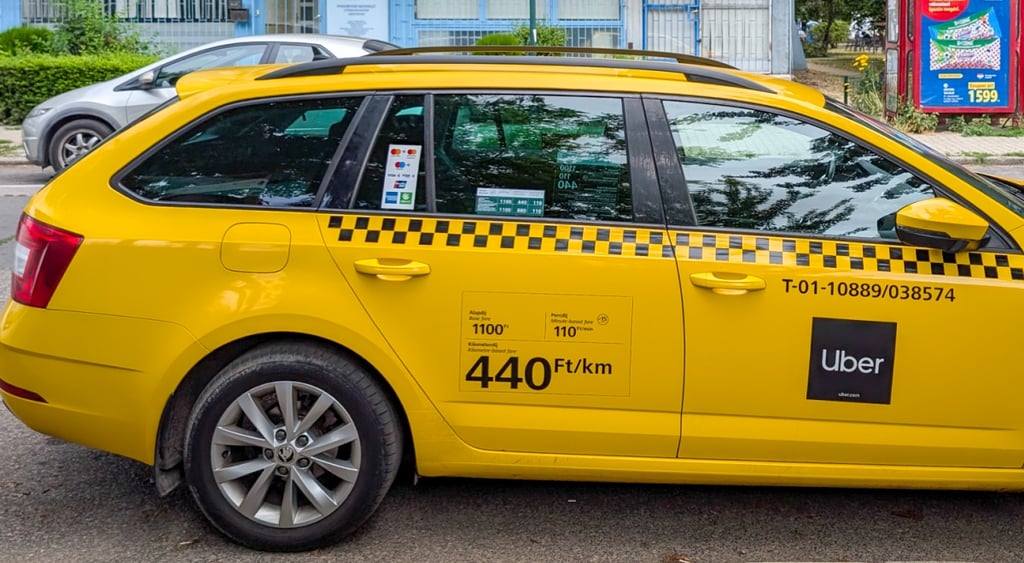 yellow taxi, uber black logo on front right door, daytime, grey car, trees, sunny day