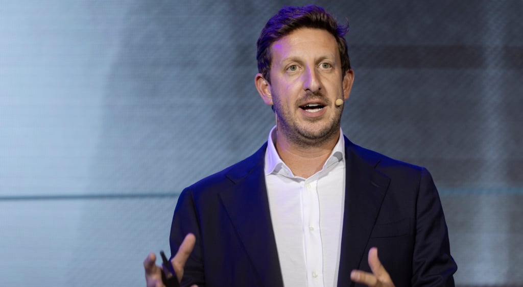 Zack Kass, middle age white male, white collar shirt, no tie, black blazer, dark hair, big ears, speaking with mic