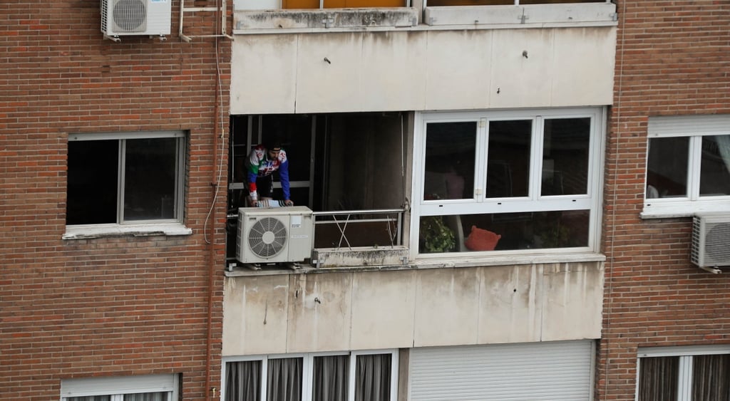 dark brown brick building, air conditioners, man wearing cyclist uniform in dark balcony, white window frames