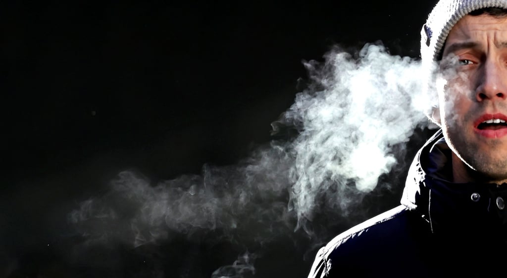 white male, grey hat, black padded jacket, pink lips, white cloud of cold breath, black background