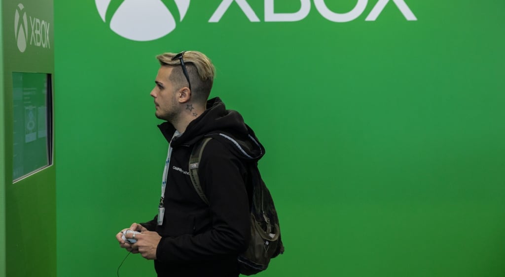 xbox white sign on green wall, white blonde male in black hoodie plays a game in front of screen