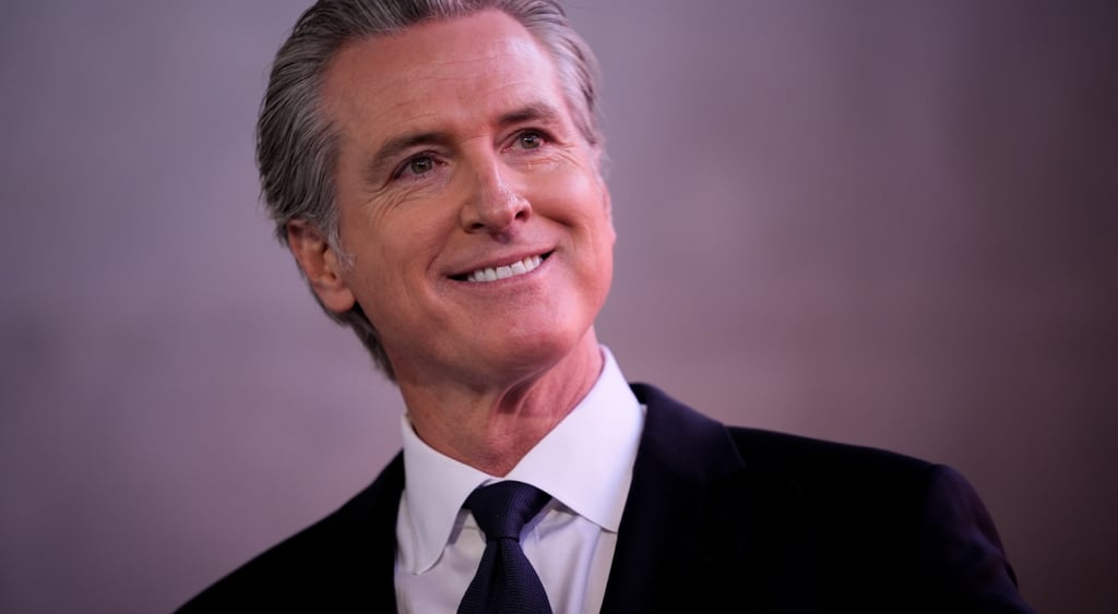 Governor Gavin Newsom white make, purple grey background, black suit and tie, white collar shirt