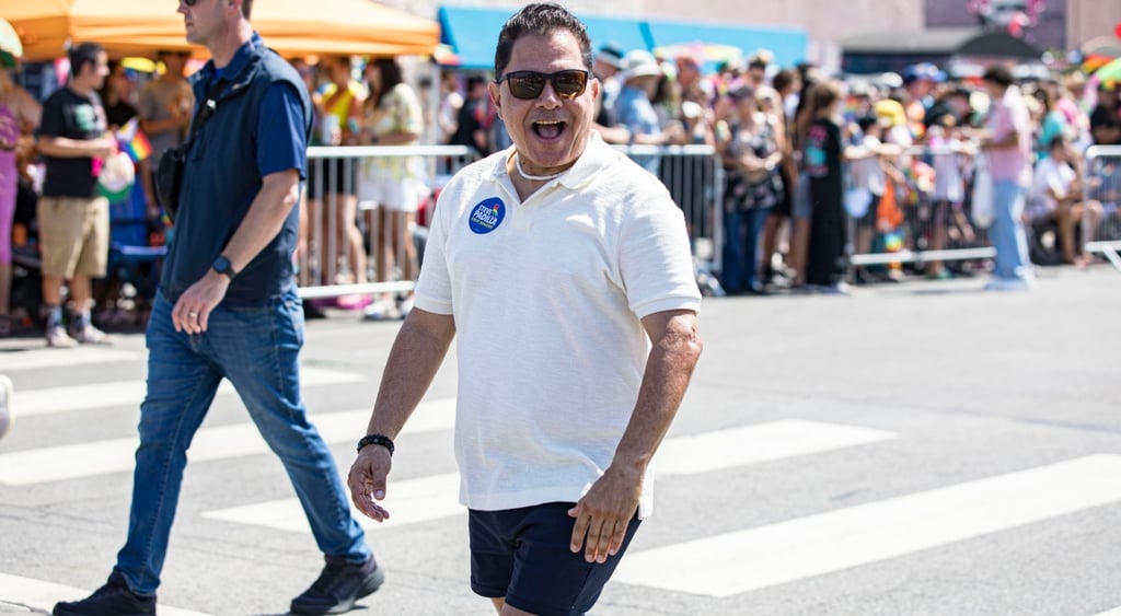 steve padilla, male in black shorts, white t-shirt, blue round badge, grey pavement, pedestrian crossing