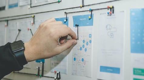 A man's hand pointing with a pin to a map hung on a wall