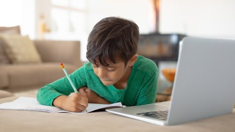 child sitting by computer and writing something with a pencil on paper