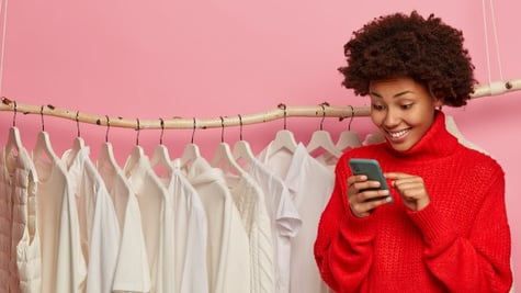Young woman looking excitedly at cellphone and a line of white clothes hooked on a rack in the backg