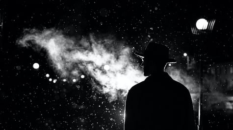 dark silhouette of a man in a hat in the rain on a night street in a city in the style of Noir