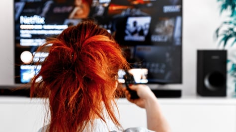 Woman sitting in her living room Holding a TV remote control and displays the netflix homepage