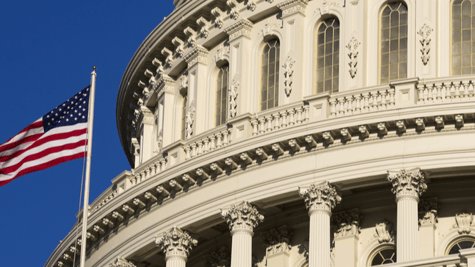 Part of the Capitol Hill building with the US flag flying on a sunny day.