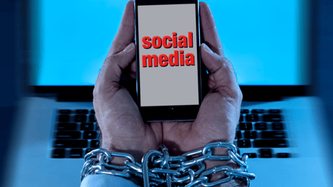Chained hands struggle to use social media on mobile phone, with computer in background