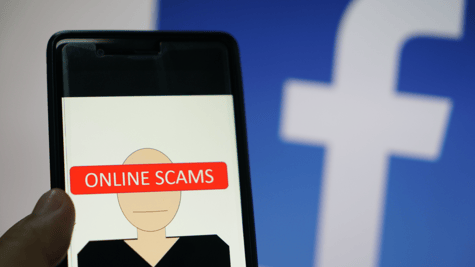 Mobile phone with scam written on it and FB logo in backdrop