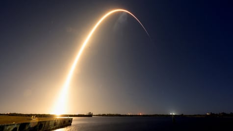 Rocket launches, leaves a yellow light trail in violet sky above water