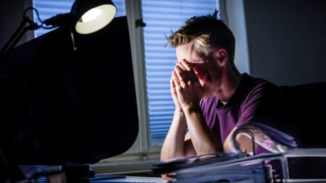 A stressed out office worker clutching his head at night, next to a bright lamp.