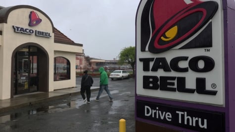 A taco bell drive thru sign and some customers approaching the restaurant