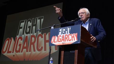 Bernie Sanders speaking on the podium among slogans reading "Fight Oligarchy"