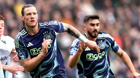 two football players run, dark blue tops, ajax logo, crowded stadium in the back, long blond hair