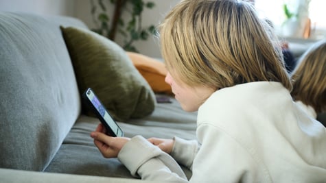 blonde boy, shoulder length hair, scrolls smartphone, white sweatshirt, grey sofa