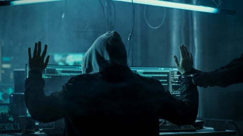 dark web hacker in hoodie, arms up, left arm handcuffed, arrest, dark blue light room