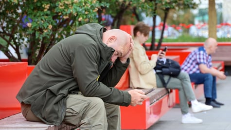 bald man, scrolls phone, red bench, dark green jacket, green trees