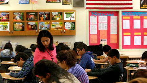 classroom, teacher in pink top, isa flag, kids leaning on tables, 