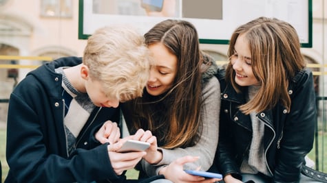 two teen girls, brunettes hold smartphones, one blonde teen boy, wait in bus stop, laugh