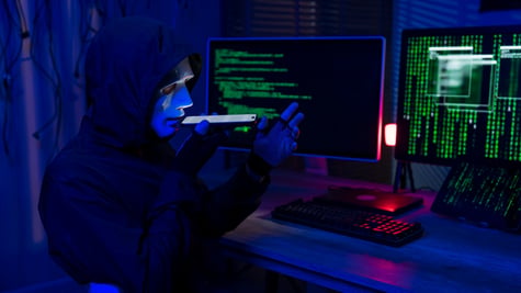 hacker, black hoody, white plastic mask on face, smartphone, two screens with green numbers, code