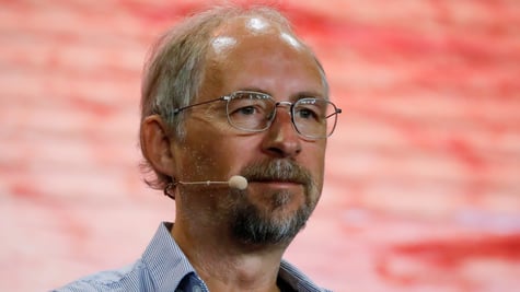 Adam Back, grey hair, tanned white skin, glasses, blue collar shirt, pink background