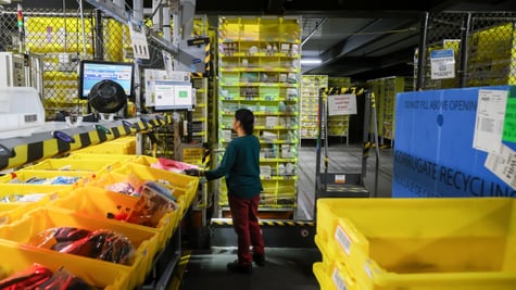 Female worker, dark hair, Amazon fulfillment center, yellow parcel boxes, fence, dark red throusers