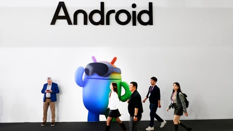 three asians walk pass blue android mascot, white wall, one man leaning to wall, Android black sign
