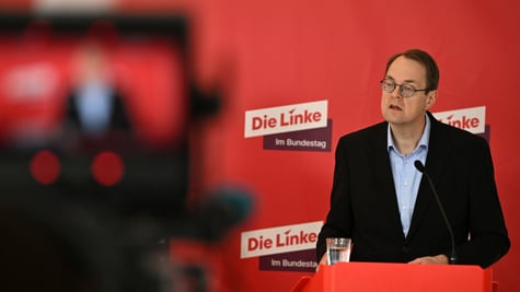 Soeren Pellmann, man in black suit, light blue shirt, no tie, collar unbuttoned, red photo wall